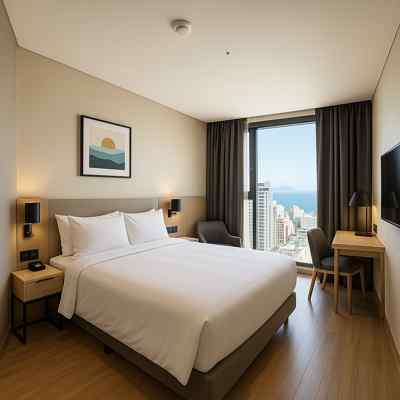 A modern ocean-view guest room at Busan Hotel L7 Haeundae by Lotte Hotel, featuring a queen-sized bed, minimalist furniture, and a city and beach view through a large window.