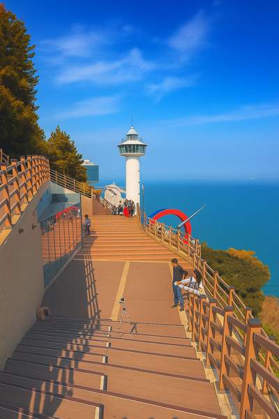 A scenic view of Taejongdae Resort's lighthouse and stairs with a deep blue sea and slight autumn foliage under a clear sky
