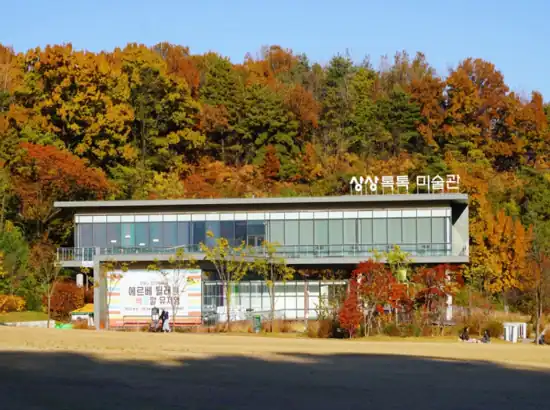 Autumn view of the Sangsang Toktok Art Museum in Buk Seoul Dream Forest travel, surrounded by colorful fall trees and open lawn space.