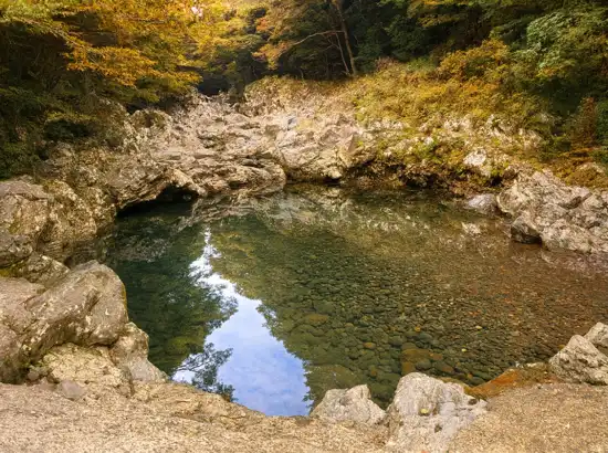 Clear autumn pool along the Hallasan Dullegil Section 3 Forest Healing Trail, reflecting trees and rocks in a calm mountain stream.
