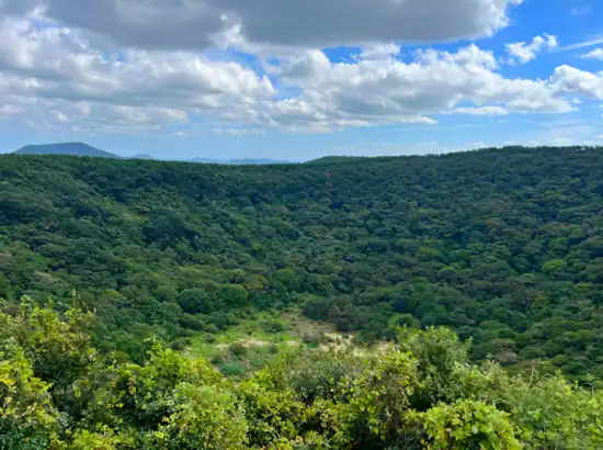 A lush green crater view along the Jeju Sangumburi Travel Course under a blue sky with scattered clouds.
