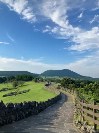 A scenic walkway along the Jeju Sangumburi Travel Course with stone walls, green fields, and a clear blue sky.
