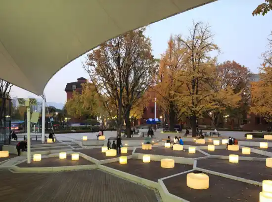 Evening view of the central plaza at Marronnier Park Seoul, with warm glowing lights, autumn trees, and people relaxing under the canopy.