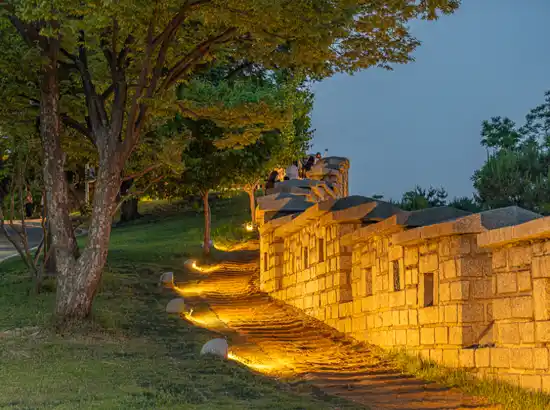 Night view of the Naksan Fortress Wall near Marronnier Park Seoul, with warm pathway lights, stone walls, and trees along the hillside.