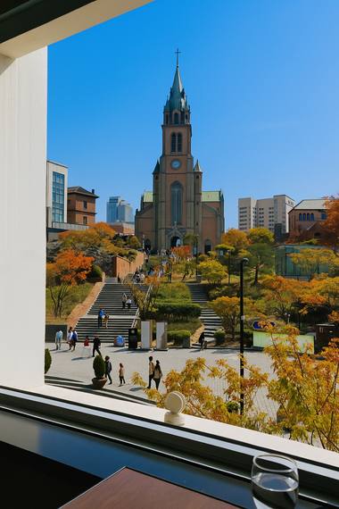 Autumn view of Myeongdong Cathedral seen from inside Pines Cafe in Seoul, with people walking across the courtyard.