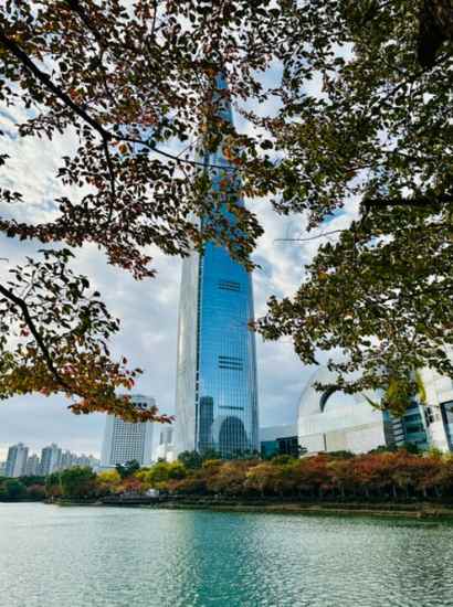 Autumn scene along the Seokchon Lake walking course, featuring Lotte Tower rising above the trees and calm turquoise water under a bright sky.