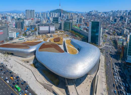 Aerial view of Dongdaemun Design Plaza (DDP) in Seoul with a clear blue sky, showing the building’s futuristic silver curves surrounded by city streets and traffic — part of the Seoul Food & Seoul Café day trip itinerary