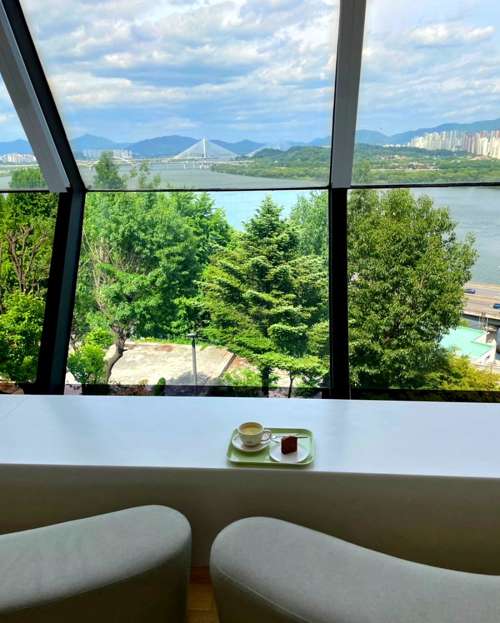 A calm café at the Grand Walkerhill Seoul with a panoramic Han River view, featuring a cup of coffee and dessert by the window.
