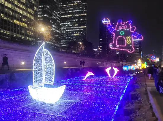 Illuminated sailboat and colorful light installations displayed over Cheonggyecheon during the 2025 Seoul Lantern Festival at night, with visitors walking along the streamside path.