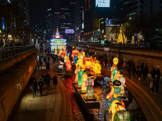 Colorful traditional lantern figures lined over water along Cheonggyecheon walkway during the 2025 Seoul Lantern Festival at night, with visitors walking beside the illuminated stream.