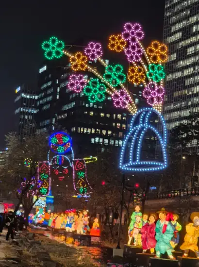 Flower-shaped lantern light installations displayed along Cheonggyecheon during the 2025 Seoul Lantern Festival at night, with illuminated buildings and visitors walking nearby.