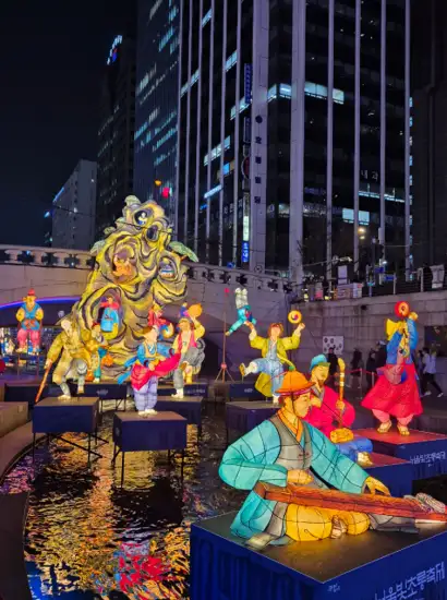 Traditional illuminated sculptures displayed over the water at the 2025 Seoul Lantern Festival near Cheonggyecheon, surrounded by visitors and high-rise buildings at night.