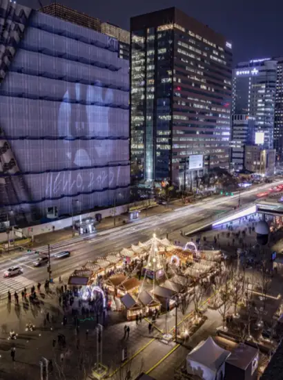 Night view of Gwanghwamun Market in Seoul during the Winter Festival, with illuminated booths, Christmas tree, and city buildings in the background.