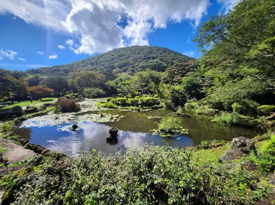 Scenic garden pond with mountain views near Jeju Jeolmul Natural Recreation Forest, reflecting blue sky and clouds.

