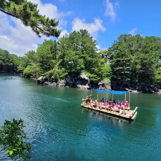 Traditional raft ride at Soesokkak Estuary in the Jeju Seogwipo travel course, with clear emerald water and forest cliffs.