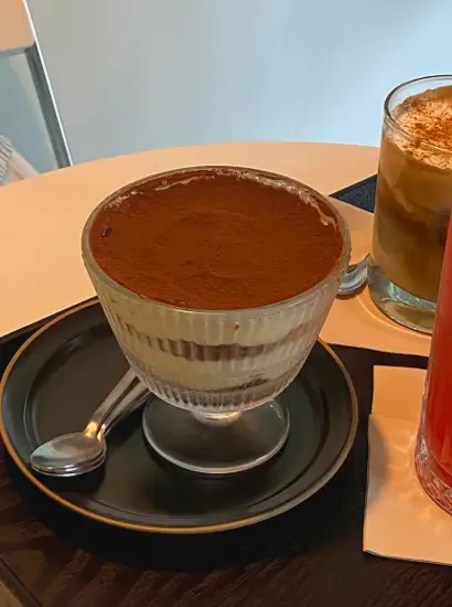 Tiramisu dessert served at a cafe near Seoul Light DDP, displayed in a clear glass bowl on a black plate with iced coffee beside it.
