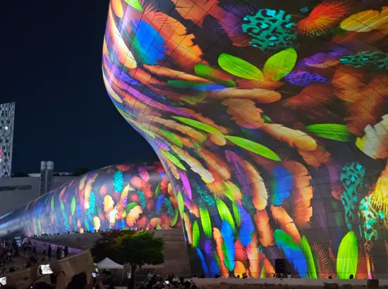 Colorful media facade projected on the exterior of Seoul Light DDP at night, showing vibrant light patterns with visitors watching outdoors.
