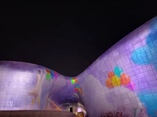 Night performance projection displayed on the curved exterior of Seoul Light DDP with colorful illumination and dancing visuals at Dongdaemun Design Plaza.
