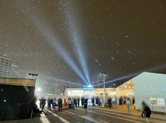 Snow falling over the Seoul Plaza Ice Rink at night with visitors waiting and winter lights in the background