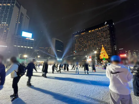People skating at the Seoul Plaza Ice Rink at night with tall buildings and a Christmas tree in the background
