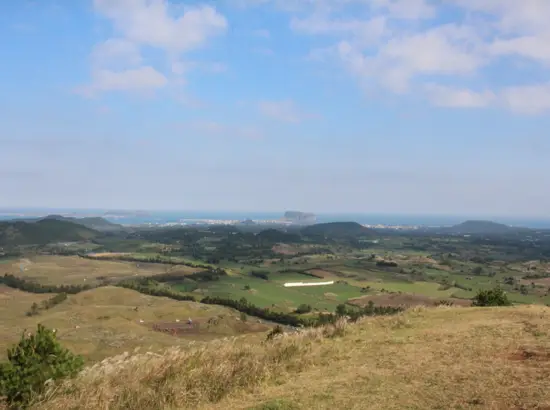 Panoramic view of Yongnuni Oreum overlooking Jeju’s eastern fields and Seongsan Ilchulbong under a clear sky.
