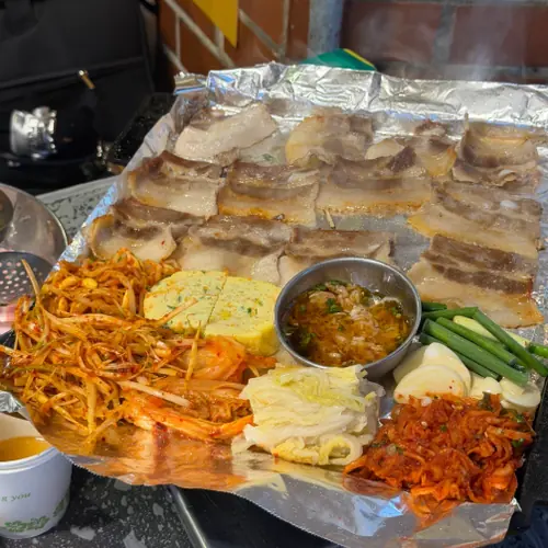 Grilled pork belly slices on a foil-lined tray with kimchi, scallion salad, green onions, dipping sauce, and side dishes.
